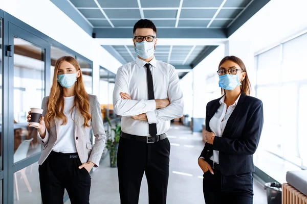 Kendine güvenen genç iş adamı lideri yüzünde tıbbi koruyucu bir maske takıyor ve arka planda bir grup meslektaşı bir ofis binasında duruyor.