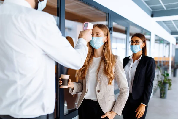 man with a non-contact thermometer measures the temperature of employees in protective masks at the entrance to a modern office, coronavirus