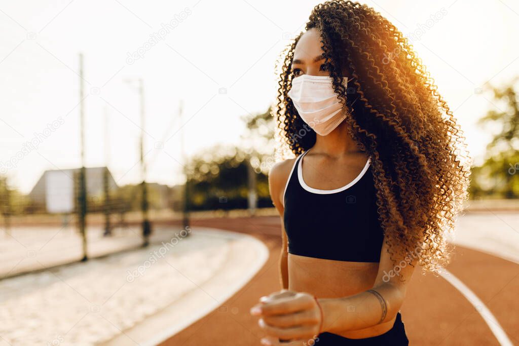 Beautiful athletic african american woman in medical protective mask on her face, jogging outdoors, fitness, people and healthy lifestyle
