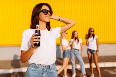Güneşli bir yaz gününde, arka planda alışveriş merkezinin yanındaki kız arkadaşlarıyla içki içen mutlu genç bir kadın.