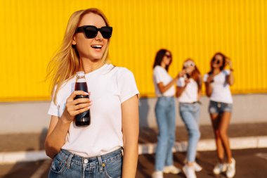 Güneş gözlüklü mutlu genç bir kadın güneşli bir yaz gününde arka planda kız arkadaşlarıyla alışveriş merkezinin yakınında içki içer.