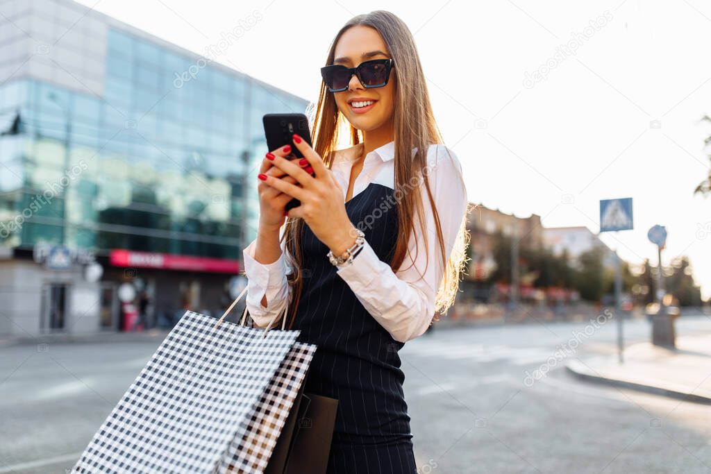 Fashionable young woman in sunglasses and a dress, with shopping bags, uses a smartphone in the city near a shopping center, shopping concept, black friday