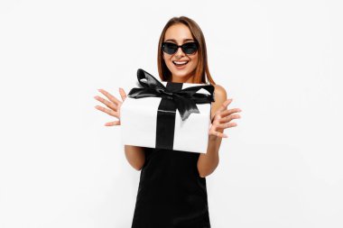 Güneş gözlüklü, zarif siyah elbiseli, beyaz arka planda hediye kutusu olan mutlu genç bir kadın. Alışveriş, tatil, Kara Cuma.