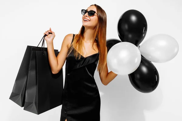 Excited young woman in black elegant dress, with balloons and shopping bags, on white background, Concept of sale, shopping, Black Friday