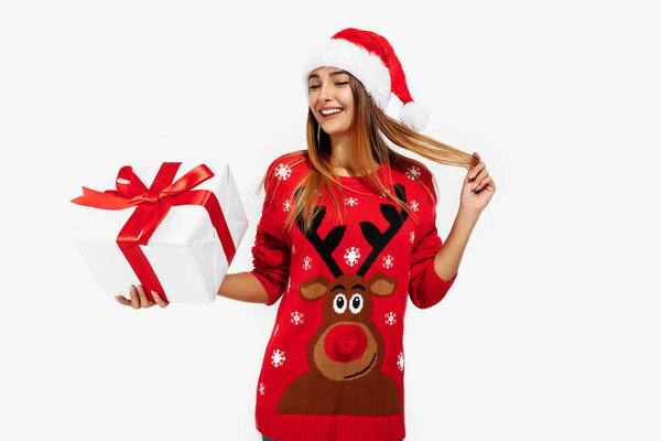 happy young woman in red Christmas sweater and Santa Claus hat, with a large gift box on a white background