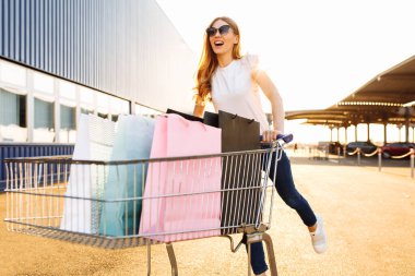 Güneş gözlüklü, alışveriş çantalı ve alışveriş arabalı güzel bir kadın güneşli bir günde şehrin arka planında bir alışveriş merkezinin önünde yürüyor.