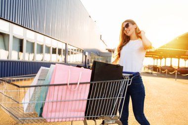 Alışveriş yapmaktan hoşlanan neşeli genç bir kadın: alışveriş merkezinin yakınında alışveriş arabasıyla çanta taşıyor ve koşuyor.