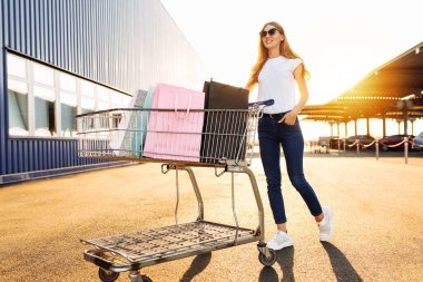 Alışveriş yapmaktan hoşlanan neşeli genç bir kadın: alışveriş merkezinin yakınında alışveriş arabasıyla çanta taşıyor ve koşuyor.