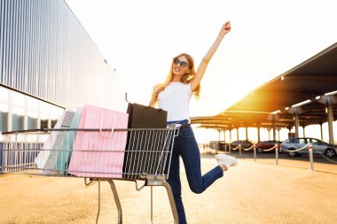 Alışveriş yapmaktan hoşlanan neşeli genç bir kadın: alışveriş merkezinin yakınında alışveriş arabasıyla çanta taşıyor ve koşuyor.