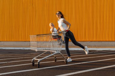 Mutlu, çocuklu genç bir kadın alışveriş merkezinin sarı duvarında alışveriş arabasıyla, aile alışverişiyle