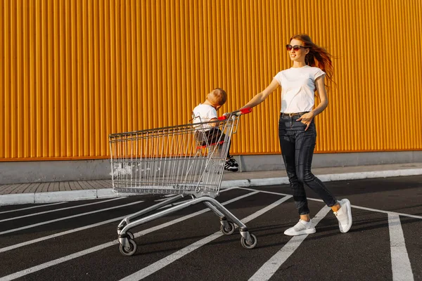 妈妈和一个小孩坐在街上的购物车里 靠着购物中心的一面黄色的墙购物 — 图库照片