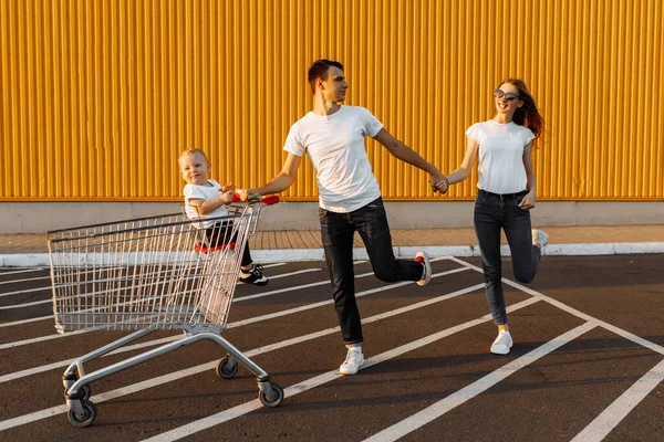 幸せな家族は子供を持つ購入 夫と妻はショッピングセンターの黄色の壁の背景を背景に ショッピングカートで楽しく走り回る — ストック写真