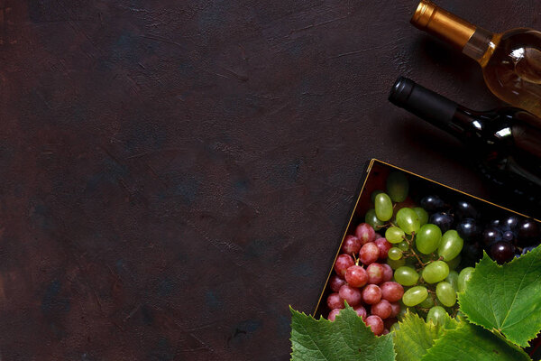 Red, green and pink grapes with leaves in metal box and two bottles of wine, white and red. Top view, close up on dark vintage background