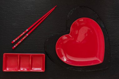 Top view of red heart shape plate on black stone board with chopsticks and saucer on black stone background clipart