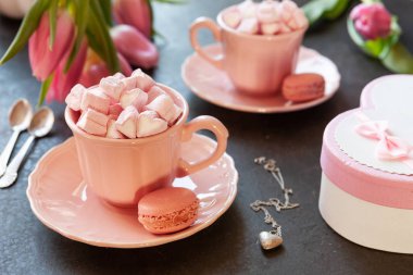 Two pink cups with pink heart shaped marshmallows with macaroons, pink tulips,  gift box and silver heart shape pendant on chain. Romantic breakfast for two. Close-up, selective focus. clipart