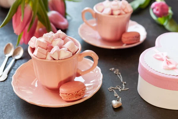 Two pink cups with pink heart shaped marshmallows with macaroons, pink tulips,  gift box and silver heart shape pendant on chain. Romantic breakfast for two. Close-up, selective focus.