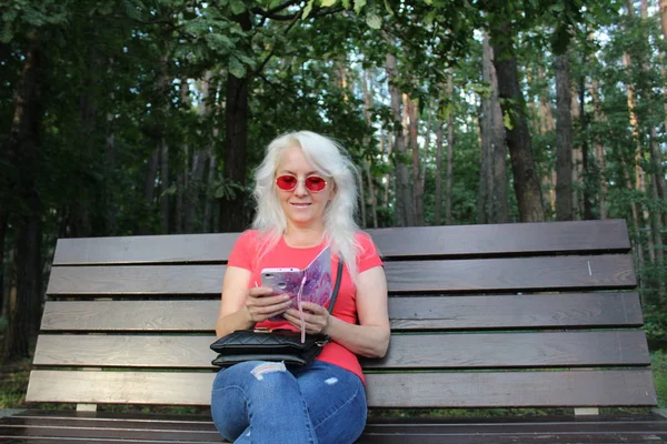 blonde girl on a Park bench / photo of a girl in the Park.a beautiful woman sitting on a bench.the blonde is resting,looking at the smartphone.Park with trees in summer.