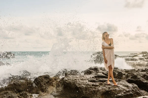 石で衝突して巨大な波の前でカメラにポーズでかわいい女の子 エキゾチックなビーチ海辺の夕日や海の日の出にセクシーな女性 運リラックスやアクティブなライフ スタイル コンセプト — ストック写真