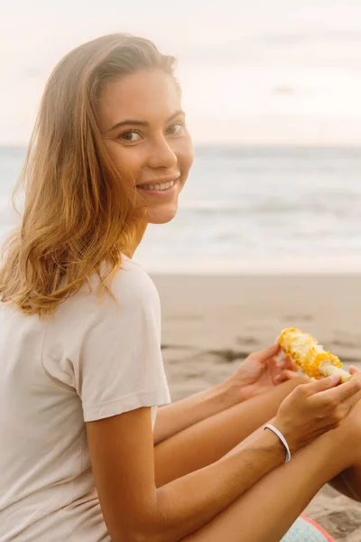 健康的な食事 ベジタリアンの内気な少女は 新鮮な有機焼きトウモロコシを食べる カメラに見えます 海ビーチの夕日や海の日の出でスポーティな女性 アクティブ ビーガン ダイエット ライフ スタイルのコンセプト — ストック写真