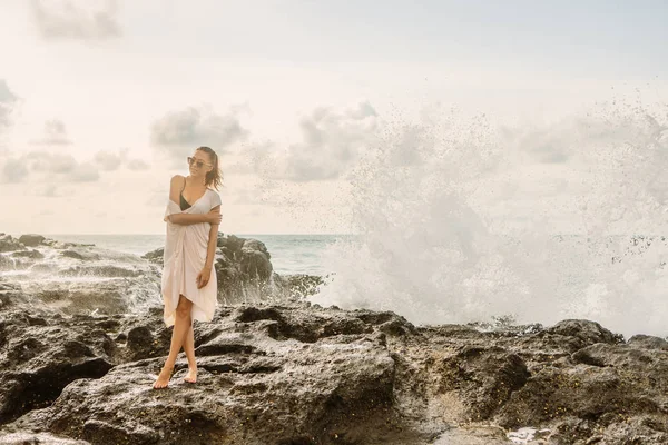 石で衝突して巨大な波の前でカメラにポーズでかわいい女の子 エキゾチックなビーチ海辺の夕日や海の日の出にセクシーな女性 サーファー リラックスやアクティブなライフ スタイル コンセプト — ストック写真
