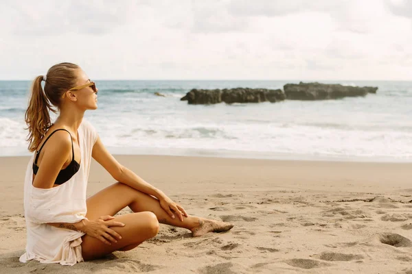 カジュアルな服を着たかなりの女性が砂の上に座り 水辺を見ています 熱帯のエキゾチックな海のビーチの夕日や海の日の出にセクシーな女性 アクティブなヨガと瞑想のライフスタイルのコンセプト — ストック写真