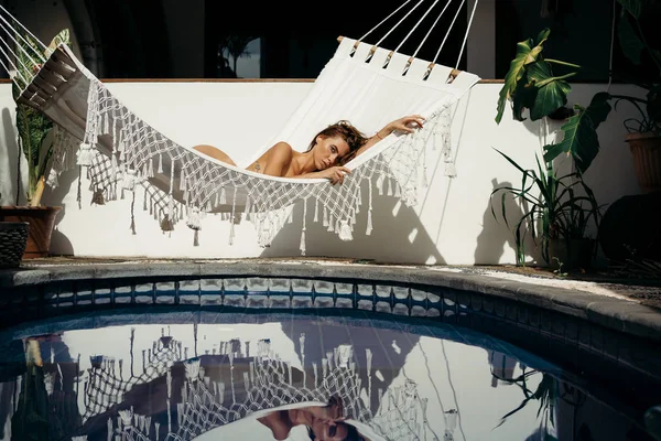 年轻漂亮的运动女孩休息和摆动在白色吊床 并反映在蓝色游泳池 热带私人住宅 愉快的妇女享受她的假期在坚硬办公室以后日常工作 — 图库照片
