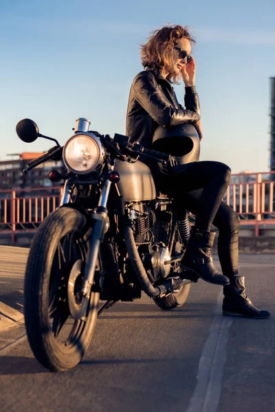 Sexy biker woman in black leather jacket sit on vintage custom caferacer motorcycle and correct sunglasses. Urban roof parking, sunset in big city. Traveling and active hipster lifestyle. Girls power.