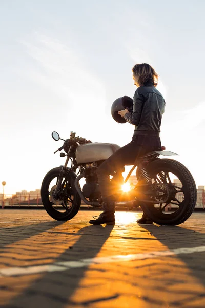 性感骑自行车的女士在黑色皮夹克和全脸头盔坐在老式定制 Caferacer 摩托车 城市屋顶停车场 日落在大城市 旅游和活跃的时髦生活方式 女孩力量 — 图库照片