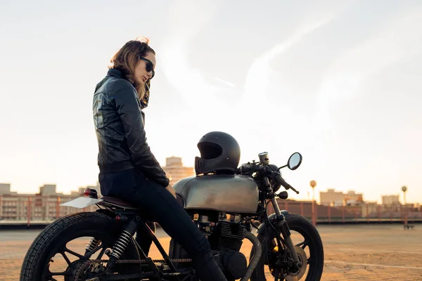性感骑自行车的女士穿着黑色皮夹克坐在老式定制 Caferacer 摩托车和触摸她的头发 城市屋顶停车场 日落在大城市 旅游和活跃的时髦生活方式 女孩力量 — 图库照片
