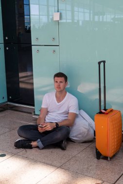 Adam havaalanında yerde oturuyor. Beyaz tişörtlü esmer.