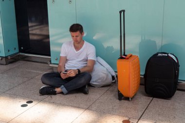 Adam havaalanında yerde oturuyor. Beyaz tişörtlü esmer.