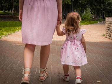 Anne ve kızı parkta yürüyüp oynuyorlar. Elbise giymişti. Güneşli bir gün, hafta sonu şehir parkında.