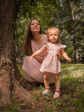 Anne ve kızı parkta yürüyüp oynuyorlar. Elbise giymişti. Güneşli bir gün, hafta sonu şehir parkında.