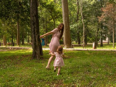 Anne ve kızı parkta yürüyüp oynuyorlar. Elbise giymişti. Güneşli bir gün, hafta sonu şehir parkında.