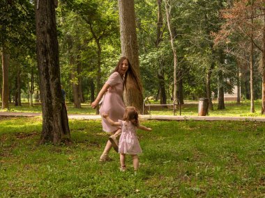 Anne ve kızı parkta yürüyüp oynuyorlar. Elbise giymişti. Güneşli bir gün, hafta sonu şehir parkında.