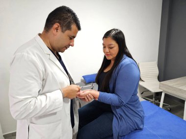 Koyu tenli erkek doktor tendondaki perküsyonu kullanarak refleks çekiçle Latin kadın nörolojik muayeneye katılıyor.