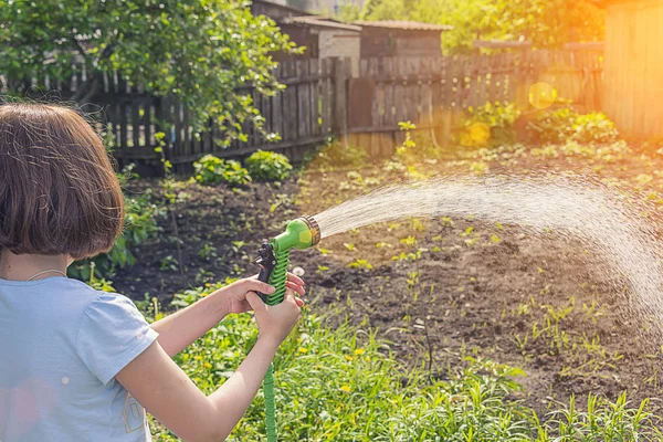 girl in a blue T-shirt watering the garden with water from a green hose in sunny weather
