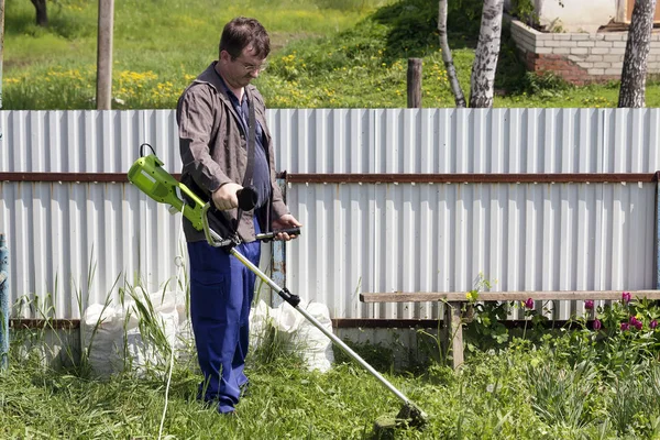 a man in glasses with a trimer mows the grass in the courtyard of a house in the village, brings order to the territory of the country