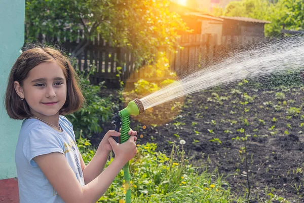 girl in a blue T-shirt watering the garden with water from a green hose in sunny weather