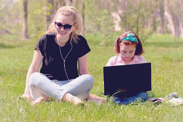 Mom and daughter resting on nature with laptop sitting on green grass in sunny weather