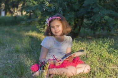 küçük kız bir albümdeki kalemler ile çim doğada bir parkta otururken çizer