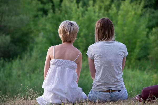 two lesbians are sitting on a hill, outdoors leaning against each other