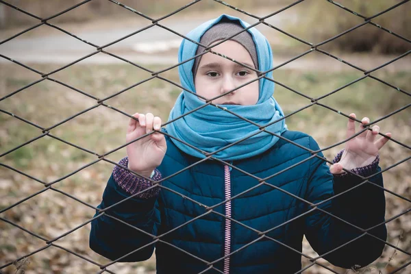 a refugee girl in a scarf standing behind bars