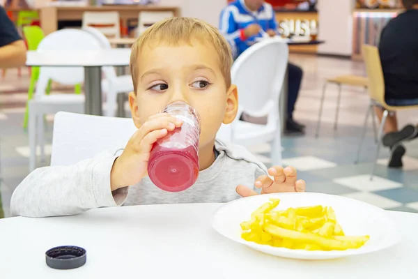 Little child eats french fries and drinks juice, sitting at the table on the food court of the mall. Unhealthy food. — Stock Photo, Image
