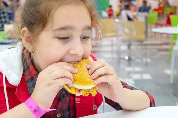 Hungry teenage girl eating a cheeseburger on the food court of the mall. Unhealthy food. — Stock Photo, Image