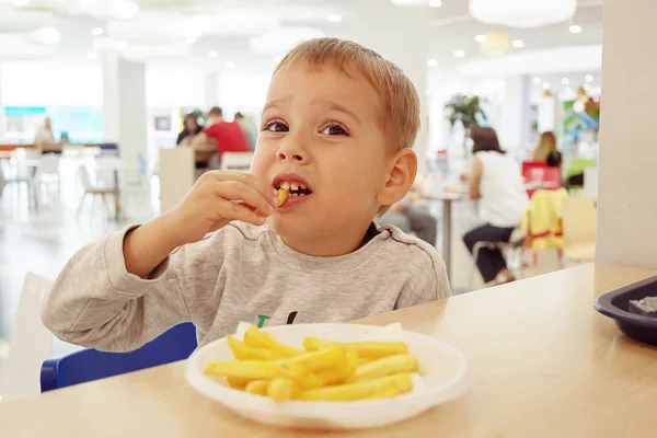 Little child eating french fries sitting at a table on the food court of the mall. Unhealthy food. — Stock Photo, Image