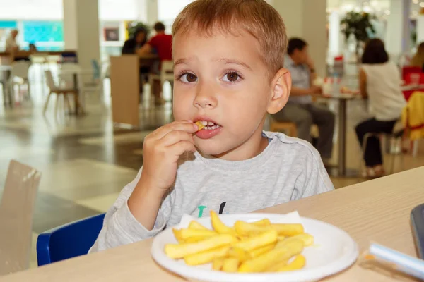 Little child eating french fries sitting at a table on the food court of the mall. Unhealthy food. — Stock Photo, Image