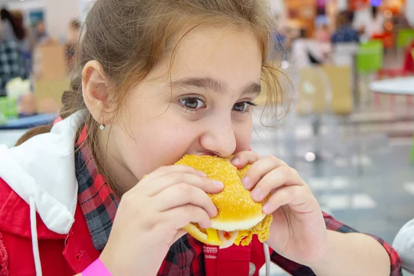 Hungry teenage girl eating a cheeseburger on the food court of the mall. Unhealthy food. — Stock Photo, Image