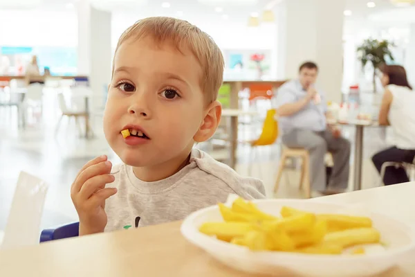 Маленький ребенок ест картошку фри, сидя за столом на фуд-корте торгового центра. Нездоровая пища . — стоковое фото