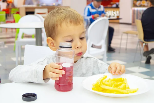 Little child eats french fries and drinks juice, sitting at the table on the food court of the mall. Unhealthy food. — Stock Photo, Image